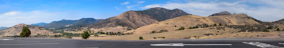 [Approximately six images stitched together with the pavement (and big white directional arrow) in the foreground and near and far mounds of land. Most of the far mountains are tree covered but the nearer mounds are covered with beige vegetation.]